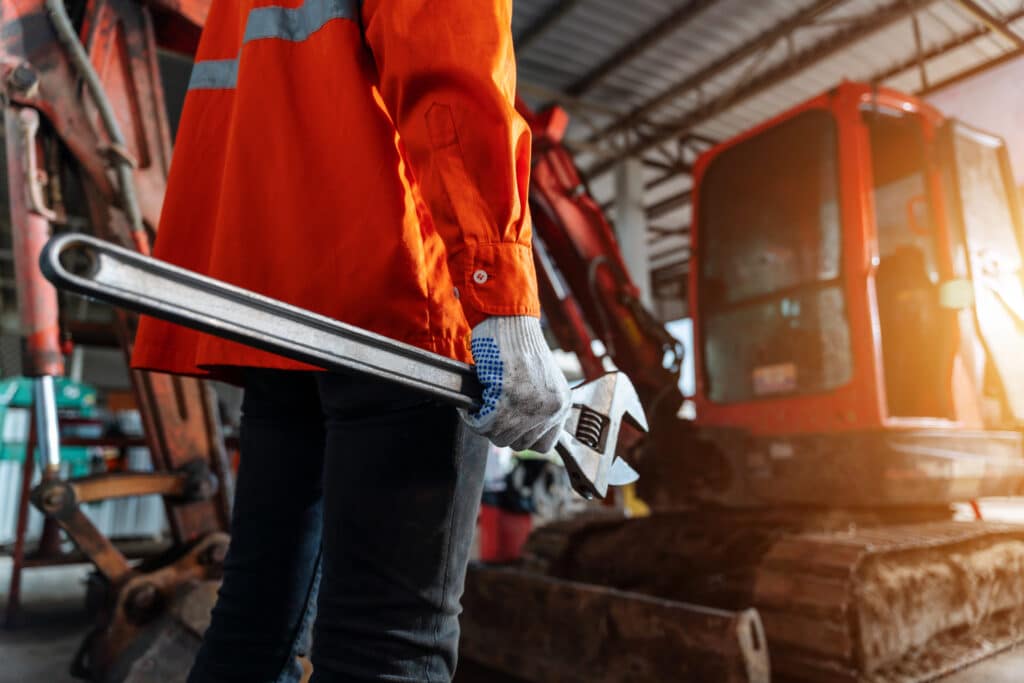 En arbejder der holder en stor skiftenøgle og står foran en gravemaskine i en industrihal eller værksted.